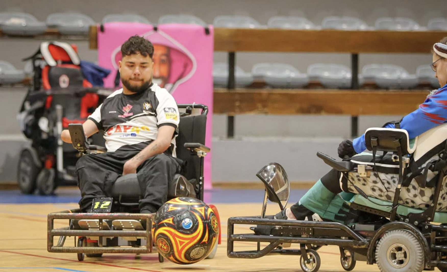 An individual seated in a dark green power wheelchair on a wooden court, with the front wheel in motion and transferring d...